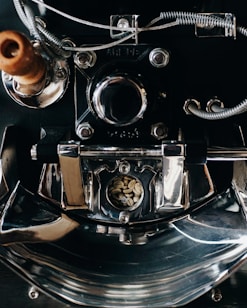 A close-up view of a metallic coffee roasting machine, highlighting its shiny, reflective surfaces and intricate components. The center features a small window displaying raw, unroasted coffee beans. Various metal coils, screws, and pipes are visible, adding complexity to the structure.