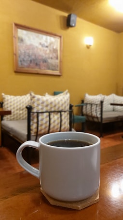 A white ceramic mug filled with coffee is placed on a wooden table in a cozy room. The background features a couple of cushioned seats with patterned pillows, set against a mustard-colored wall adorned with a painting.