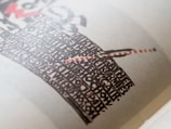A close-up of a printed page featuring a bold, artistic design with black and red text forming a shape reminiscent of a sword or a cross on an off-white background. The text style is decorative and dense, creating a visually striking pattern.