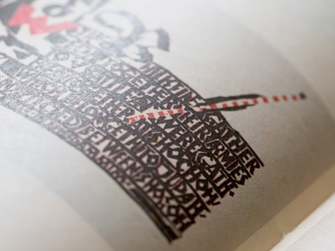 A close-up of a printed page featuring a bold, artistic design with black and red text forming a shape reminiscent of a sword or a cross on an off-white background. The text style is decorative and dense, creating a visually striking pattern.