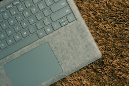 A close-up view of a laptop with a grey textured surface, positioned on a grassy background. The keyboard and touchpad are clearly visible, highlighting the device’s sleek and modern design.