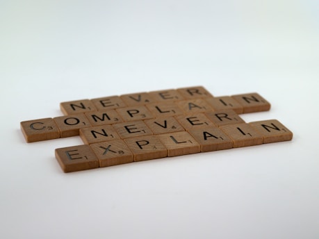Wooden letter tiles arranged to form the phrase 'Never Complain, Never Explain.' The tiles resemble those used in word games and are placed in a staggered formation on a plain surface.