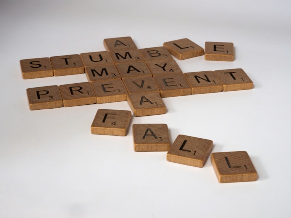 Wooden Scrabble tiles are arranged on a surface forming intersecting words such as 'STUMBLE,' 'PREVENT,' 'FALL,' and 'MAY.'