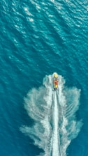 An aerial view captures a jet ski speeding through calm turquoise waters, creating a dramatic wake behind it. The sunlight reflects off the surface, adding a shimmering effect to the scene.