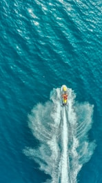 An aerial view captures a jet ski speeding through calm turquoise waters, creating a dramatic wake behind it. The sunlight reflects off the surface, adding a shimmering effect to the scene.