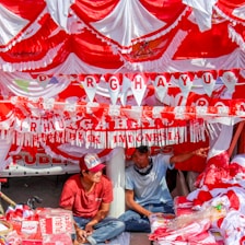 Red and white decorations hang in layers with two men seated beneath them. The banners and bunting include the word 'DIRGAHAYU' celebrating Indonesian independence. The men are surrounded by red and white items, possibly flags or merchandise.