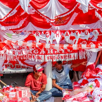 Red and white decorations hang in layers with two men seated beneath them. The banners and bunting include the word 'DIRGAHAYU' celebrating Indonesian independence. The men are surrounded by red and white items, possibly flags or merchandise.