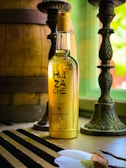 A bottle of tequila with the label 'Huizache Origen' is placed on a table near a window. The bottle is illuminated by natural light, which highlights the golden color of the liquid inside. Two ornate candlesticks and a large wooden barrel are present in the background, creating a rustic ambiance.