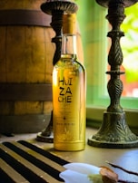 A bottle of tequila with the label 'Huizache Origen' is placed on a table near a window. The bottle is illuminated by natural light, which highlights the golden color of the liquid inside. Two ornate candlesticks and a large wooden barrel are present in the background, creating a rustic ambiance.