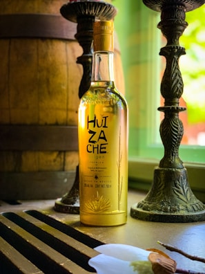 A bottle of tequila with the label 'Huizache Origen' is placed on a table near a window. The bottle is illuminated by natural light, which highlights the golden color of the liquid inside. Two ornate candlesticks and a large wooden barrel are present in the background, creating a rustic ambiance.