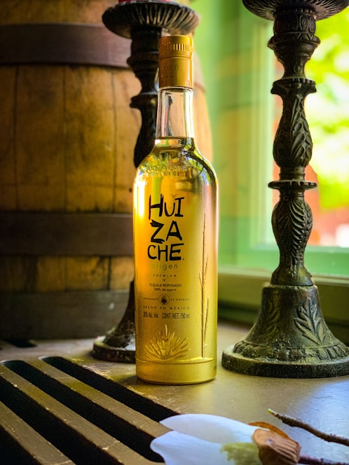 A bottle of tequila with the label 'Huizache Origen' is placed on a table near a window. The bottle is illuminated by natural light, which highlights the golden color of the liquid inside. Two ornate candlesticks and a large wooden barrel are present in the background, creating a rustic ambiance.