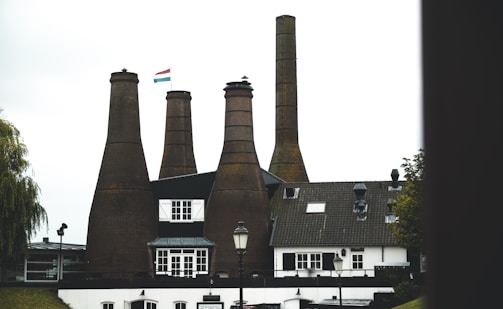 Four tall brick chimneys tower over a traditional building with white walls and dark roof, adorned with a flag featuring red, white, and blue stripes. The scene includes a streetlamp and surrounding greenery.
