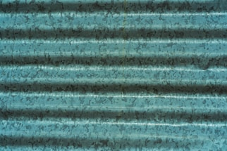 A corrugated metal sheet with a weathered, textured surface in shades of teal and dark blue, displaying irregular patterns and shadows across the ridges.