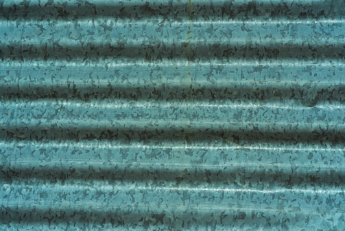A corrugated metal sheet with a weathered, textured surface in shades of teal and dark blue, displaying irregular patterns and shadows across the ridges.