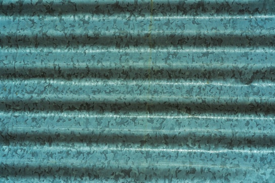 A corrugated metal sheet with a weathered, textured surface in shades of teal and dark blue, displaying irregular patterns and shadows across the ridges.