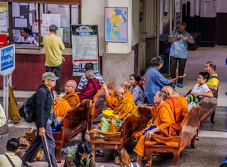 A group of Buddhist monks in orange robes are seated together in a public waiting area, engaging in conversation. Other people are also present, either sitting or walking past. The setting appears to be a busy station or terminal with informational signs and a ticket counter in the background.