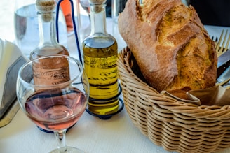 A wicker basket contains a loaf of rustic bread alongside a glass of rosé wine, and two glass bottles filled with oil and vinegar. The setting is on a table covered with a white tablecloth, and cutlery is partially visible next to the basket.