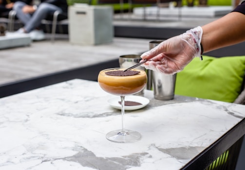 A gloved hand is delicately placing a chocolate garnish on top of a creamy cocktail in a martini glass. The setting is a modern outdoor space with a marble table, green cushions, and stainless steel shakers in the background.