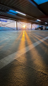 Sunlight streams through metal bars, casting long shadows across the textured floor of a parking garage. A car is parked on the right, and fluorescent lights hang from the ceiling. A sign indicating 'Ausfahrt/Exit' is visible on the left.