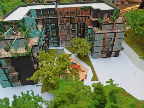 A scale model of a modern residential building complex featuring multiple stories with balconies adorned with plants. The structure includes varied textures and colors on the facade, such as teal, black, and brick. In the foreground, a small courtyard with children's playground equipment is surrounded by miniature trees and pathways, giving a sense of a green urban environment.
