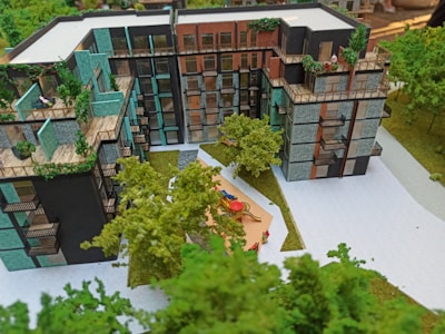 A scale model of a modern residential building complex featuring multiple stories with balconies adorned with plants. The structure includes varied textures and colors on the facade, such as teal, black, and brick. In the foreground, a small courtyard with children's playground equipment is surrounded by miniature trees and pathways, giving a sense of a green urban environment.