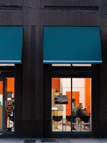 A hair salon is visible through the large glass windows of a building, with a person getting a haircut by a hairstylist inside. The salon interior features bright orange and white walls, and there is a sign on the window informing customers they can come in without an appointment.