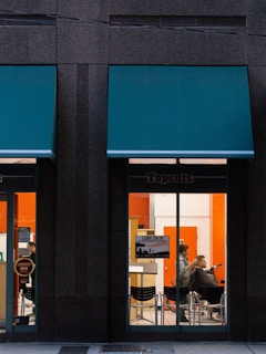 A hair salon is visible through the large glass windows of a building, with a person getting a haircut by a hairstylist inside. The salon interior features bright orange and white walls, and there is a sign on the window informing customers they can come in without an appointment.