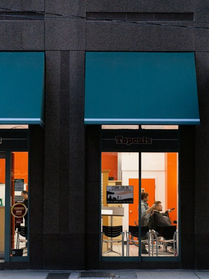 A hair salon is visible through the large glass windows of a building, with a person getting a haircut by a hairstylist inside. The salon interior features bright orange and white walls, and there is a sign on the window informing customers they can come in without an appointment.