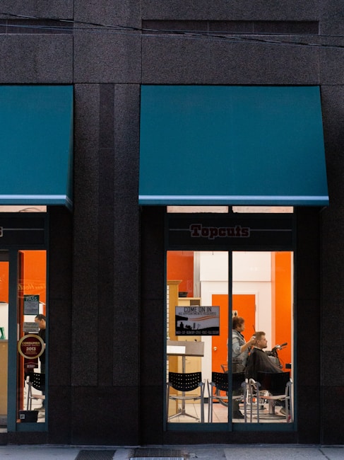 A hair salon is visible through the large glass windows of a building, with a person getting a haircut by a hairstylist inside. The salon interior features bright orange and white walls, and there is a sign on the window informing customers they can come in without an appointment.