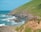 A group of people wearing helmets and outdoor gear stand on the edge of a grassy cliff overlooking a rocky coastline. The ocean waves crash against the rocks below, with a backdrop of blue skies and lush green hills.