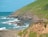 A group of people wearing helmets and outdoor gear stand on the edge of a grassy cliff overlooking a rocky coastline. The ocean waves crash against the rocks below, with a backdrop of blue skies and lush green hills.