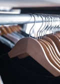 A row of natural wood hangers is aligned neatly on a metal rack, casting shadows and showing the smooth texture of the wood. The hangers hold dark-colored garments in a softly lit environment, suggesting a clothing store or wardrobe setting.