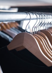 A row of natural wood hangers is aligned neatly on a metal rack, casting shadows and showing the smooth texture of the wood. The hangers hold dark-colored garments in a softly lit environment, suggesting a clothing store or wardrobe setting.