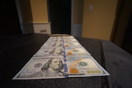 A row of U.S. hundred-dollar bills is neatly arranged on a dark surface. The focus is on the foreground bills, with details of Benjamin Franklin's portrait visible. The lighting casts soft shadows, giving the scene a slightly moody atmosphere.