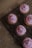 Five cupcakes with pink frosting and colorful sprinkles are arranged on a black cooling rack. The cupcakes are placed on a brown paper background with scattered sprinkles around the rack.