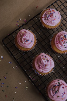 Five cupcakes with pink frosting and colorful sprinkles are arranged on a black cooling rack. The cupcakes are placed on a brown paper background with scattered sprinkles around the rack.