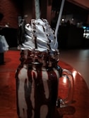 A decadent milkshake topped with whipped cream and chocolate sauce, served in a large glass jar. An Oreo cookie is placed on the side, and two straws extend from the top. The background has a warm, dimly lit ambiance, suggesting a cozy setting.