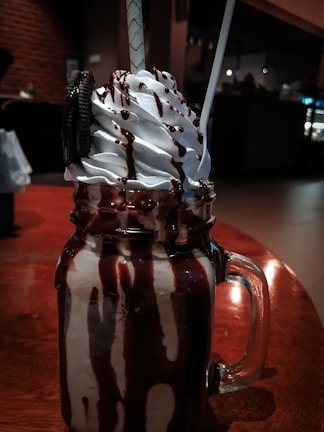 A decadent milkshake topped with whipped cream and chocolate sauce, served in a large glass jar. An Oreo cookie is placed on the side, and two straws extend from the top. The background has a warm, dimly lit ambiance, suggesting a cozy setting.