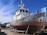 A large, weathered fishing boat is dry-docked and supported by metal stands. The hull is covered with patches of rust and peeling paint. The boat has a white cabin with windows, and various antennas and equipment are mounted on top. In the background, there are other boats and a bright blue sky with some wispy clouds.