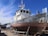 A large, weathered fishing boat is dry-docked and supported by metal stands. The hull is covered with patches of rust and peeling paint. The boat has a white cabin with windows, and various antennas and equipment are mounted on top. In the background, there are other boats and a bright blue sky with some wispy clouds.