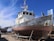 A large, weathered fishing boat is dry-docked and supported by metal stands. The hull is covered with patches of rust and peeling paint. The boat has a white cabin with windows, and various antennas and equipment are mounted on top. In the background, there are other boats and a bright blue sky with some wispy clouds.