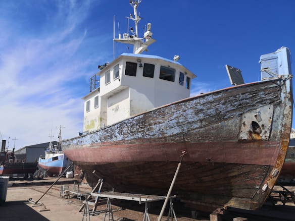 A large, weathered fishing boat is dry-docked and supported by metal stands. The hull is covered with patches of rust and peeling paint. The boat has a white cabin with windows, and various antennas and equipment are mounted on top. In the background, there are other boats and a bright blue sky with some wispy clouds.