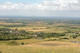 A vast rural landscape featuring a patchwork of fields and farmland stretching into the distance. The fields vary in color, indicating different crops or stages of growth. In the foreground, a small village is visible with houses nestled among trees. The sky is partly cloudy, with sunlight casting a gentle glow over the scenery.