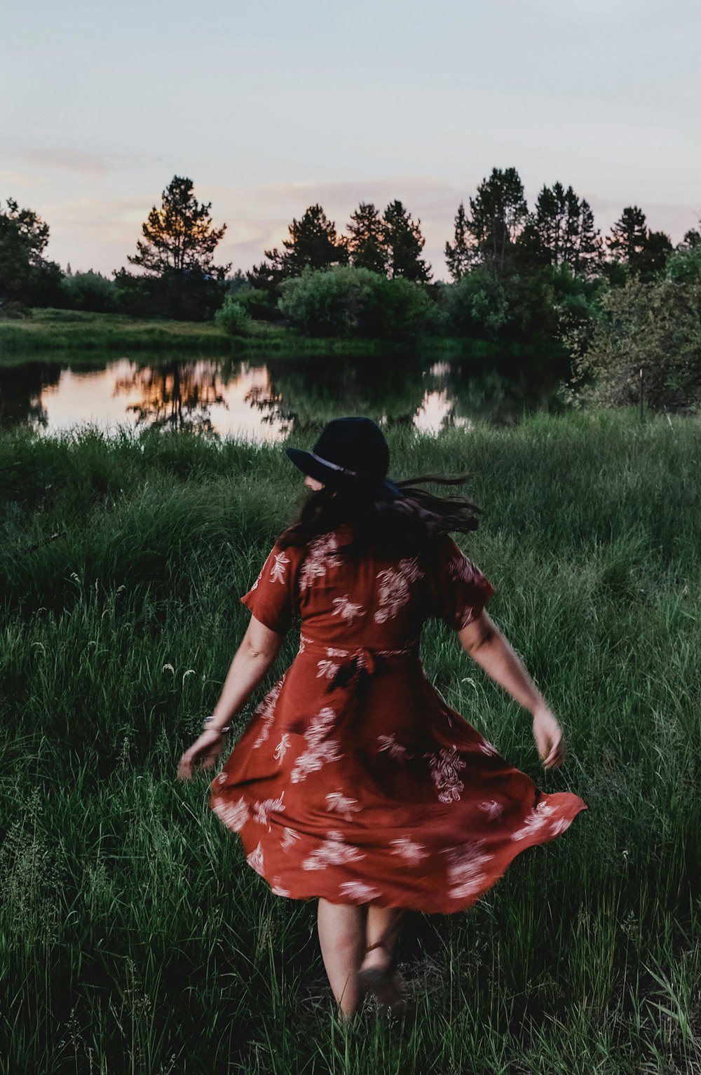 woman in red dress and black hat sitting on green grass field near lake during daytime