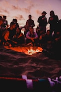A group of men gathered around a campfire on the beach at sunset.