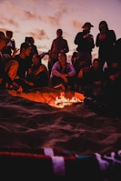 A group of diverse people gathered around a campfire sharing stories at dusk.