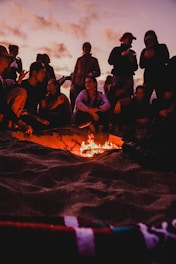 A group of people gathered around a small campfire on a beach, silhouetted against a twilight sky with scattered clouds. Some are sitting while others are standing, engaged in conversation or using their phones. The warm glow of the fire contrasts with the cool tones of the evening.