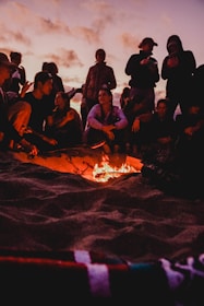 A lively beach scene with friends laughing around a campfire at sunset.