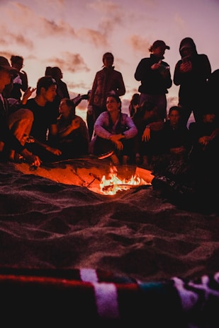 An evening camp setup by the shore with tents and a small group gathered around a fire.