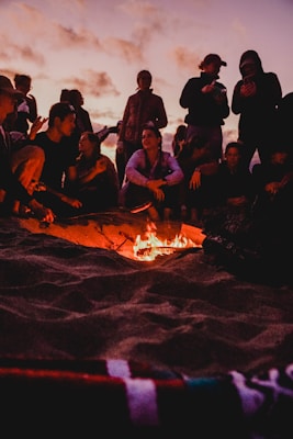 A group of people gathered around a small campfire on a beach, silhouetted against a twilight sky with scattered clouds. Some are sitting while others are standing, engaged in conversation or using their phones. The warm glow of the fire contrasts with the cool tones of the evening.